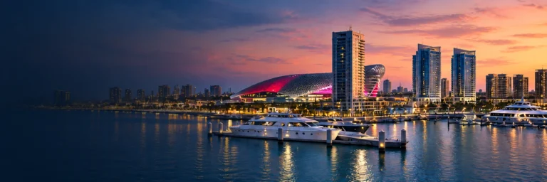 Yas Marina at dusk with yachts, waterfront towers and a lit lattice-roofed building